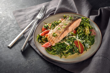 Salmon fillet roasted with a crispy skin on cream spinach vegetables with onions and tomatoes, healthy fish dish with cutlery on a dark gray napkin and slate surface, copy space