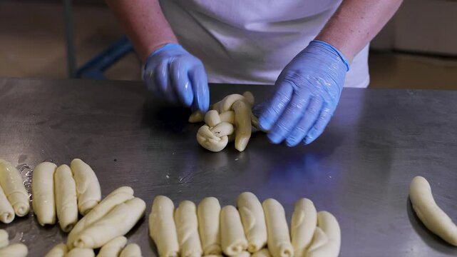 Two women with blue gloves who work in a bakery skillfully weave together three strands of dough filled with walnut paste. This detailed view captures the hands-on process of making traditional walnut