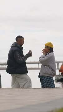 Vertical side view medium shot of young Biracial couple getting engaged standing outdoors by water on cool spring day. Guy putting engagement ring on girlfriend finger and they kissing