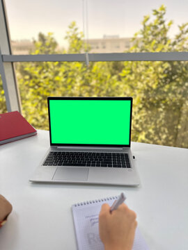 a woman watching video tutorials, making notes with a pen in an office in front of a widow with a view of tall green trees on a bright sunny day
