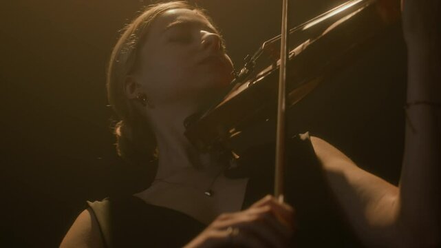 Expressive young Caucasian female music performer playing violin under projector light on dark stage