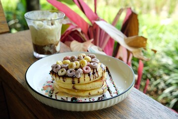 A stack of fluffy pancakes topped with colorful cereal, drizzled with syrup, and accompanied by a glass of creamy dessert. Perfect for food blogs, breakfast menus, or healthy snack promotions.