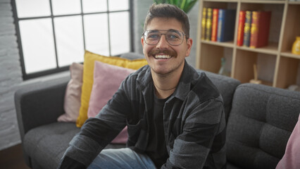 A cheerful young hispanic man with a moustache sitting on a sofa in a cozy living room, smiling at...