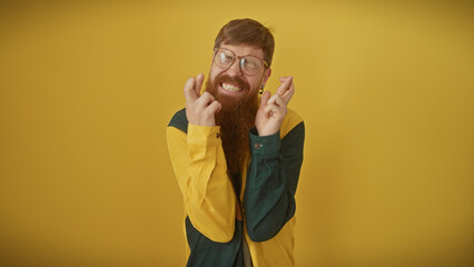 Redhead bearded man in glasses smiles with closed eyes, making fingers crossed gesture against a...