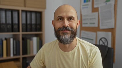 Handsome hispanic man with beard and bald head smiling in an office with shelves of binders in the...