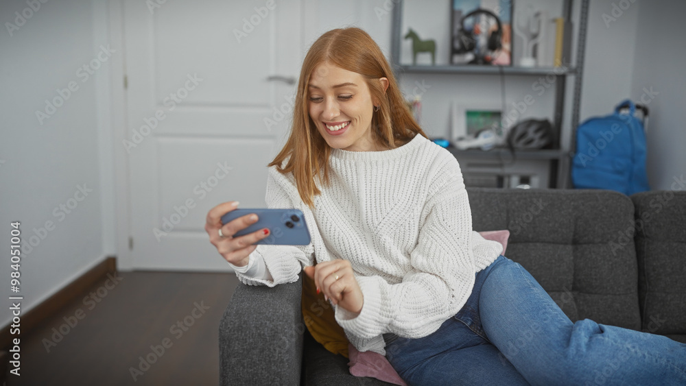 Poster A smiling young woman lounging on a couch in a cozy living room while using her smartphone.