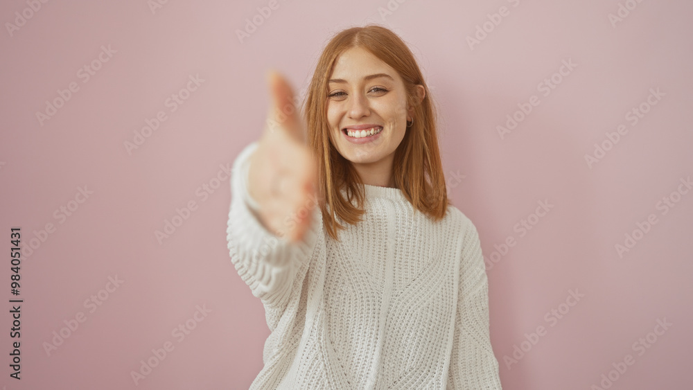 Sticker Smiling young redhead woman extends her hand in greeting against a pink background.