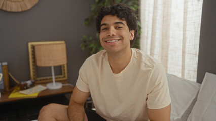 A handsome young hispanic man smiles while sitting on his bed in a cozy, well-decorated bedroom, creating a warm and inviting indoor scene at home.