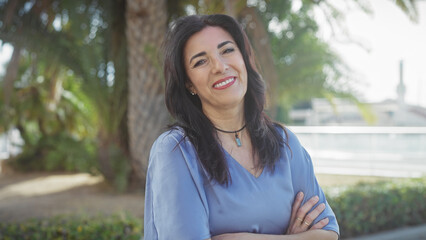 Smiling middle-aged hispanic woman with arms crossed standing outdoors in a lush park
