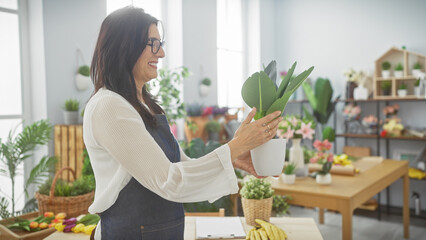 Mature hispanic woman arranges plants in sunny flower shop interior, embodying entrepreneurship and care.