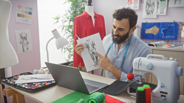 A bearded tailor in a design studio points at a sketch during a video call, with a sewing machine and fabrics nearby.