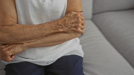 Naklejka premium An elderly woman with crossed hands sitting in a living room, highlighting the mature wrinkles and textures of her skin.
