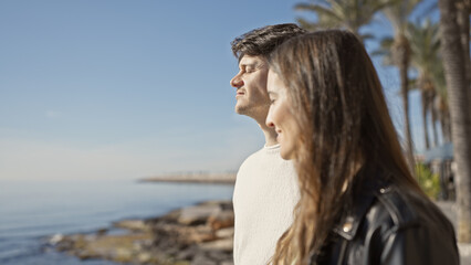 Contemplative couple enjoying serene ocean view at a rocky seaside promenade with palm trees