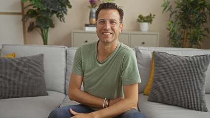 Handsome hispanic man sitting on a sofa in a cozy living room, smiling confidently towards the camera, surrounded by soft cushions and green plants, interior of a modern home