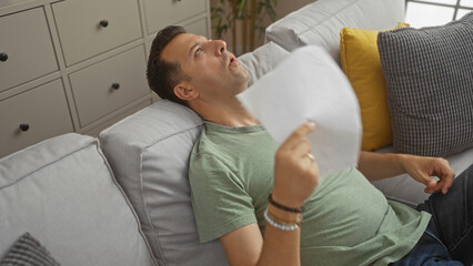 Hispanic man sits in a living room fanning himself with a paper, appearing exhausted or overheated...