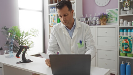 Hispanic man working in a pharmacy wearing a lab coat, focusing on a document at his laptop in a well-organized store