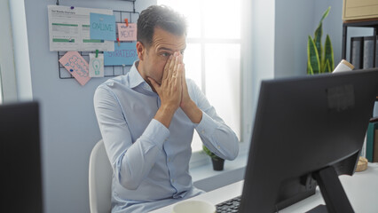 Stressed middle-aged hispanic man in office working on computer with hands on face surrounded by...