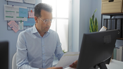 Man working in office sitting at desk with computer looking at documents surrounded by charts and papers on wall