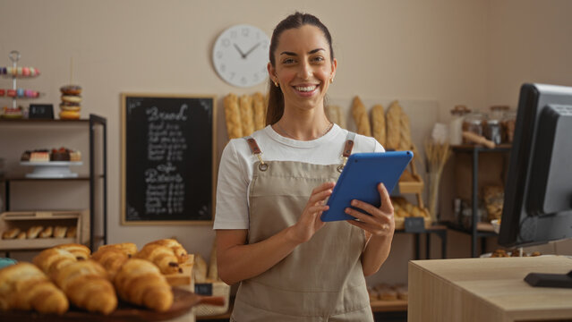 Smiling woman working in a bakery shop using a tablet surrounded by fresh pastries and breads - Powered by Adobe