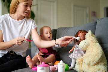 Mum and baby sitting on the sofa role-playing, feeding a doll from baby dishes.