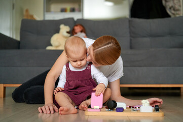 Fototapeta premium Mother in white T-shirt and grey jeans and young daughter in burgundy overalls and light blouse playing on the floor of a flat.