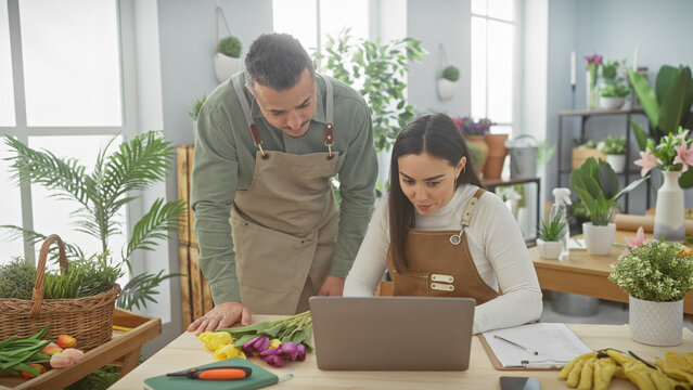 A man and woman in aprons use a laptop in a florist shop surrounded by plants and flowers, representing teamwork in a small business.