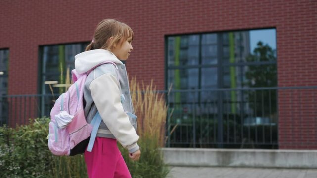 Young girl, 8 years old, is walking to school with a calm and focused expression. Kid is wearing a grey and white varsity jacket, bright pink pants, and carrying a pink backpack. Side shoot.