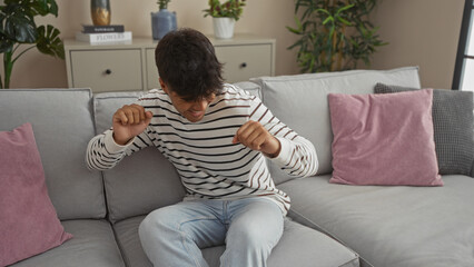 A young, handsome hispanic man is sitting and smiling on a comfortable couch in a cozy living room,...