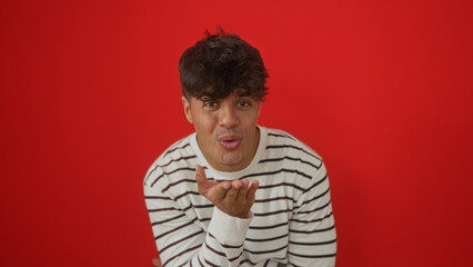 Young, attractive hispanic man wearing a striped shirt blowing a kiss, isolated against a vibrant red background.