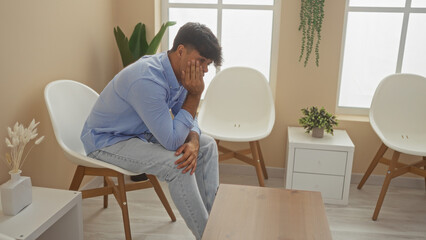 Hispanic man sitting in a waiting room with a worried expression, surrounded by modern furniture and indoor plants.