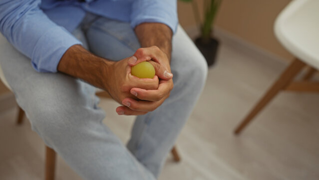 A young hispanic man sits indoors in a waiting room, gripping a stress ball tightly with both hands, wearing light clothing and appearing thoughtful.