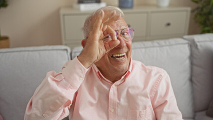 A cheerful elderly caucasian man in glasses and a pink shirt is sitting in a living room, making an ok gesture with his hand, displaying a joyful expression in a home interior setting.