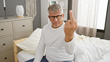 Middle-aged man in glasses sitting on a bed indoors, making a rude hand gesture in a bedroom setting