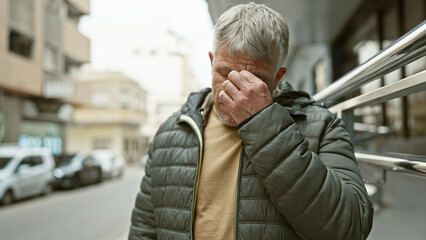 A grey-haired man appears distressed on a city street, covering his face with his hand.