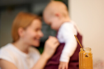 Baby food jar in the foreground, spoon inside, mum and baby in the background in out of focus. Mum and girl in white t-shirts.