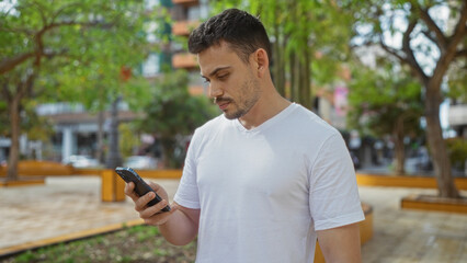 Obraz premium A handsome young hispanic man with a beard looks at his smartphone while standing in an urban park in a city setting on a sunny day.