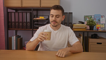 Handsome young hispanic man enjoying a coffee while sitting in an organized office with shelves,...