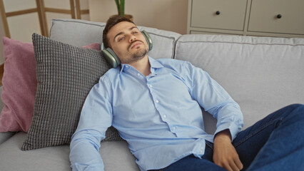 Fototapeta premium Relaxed young hispanic man wearing headphones and a blue shirt lounging on a comfortable couch in a modern living room with pillows.