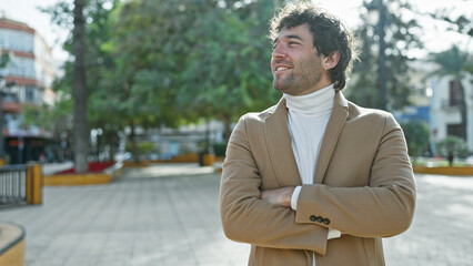 Handsome hispanic man with a beard, smiling in an outdoor park setting, standing with arms crossed.