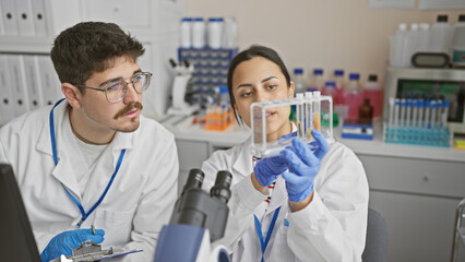 A man and woman in lab coats examine test tubes together in a scientific laboratory indoor setting.