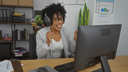 Young african american woman celebrating success in modern office environment with headphones on at her workplace desk.