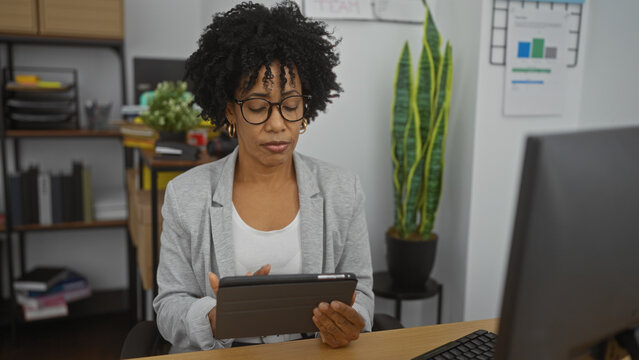 African american woman with curly hair using a tablet in an indoor office with plants and shelves in the background, focusing deeply on her work.