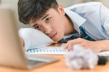 Obraz premium A young man appears bored and tired while working on a laptop, resting his head on his arm and surrounded by documents.