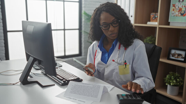 A young african american woman in a clinic room, dressed as a doctor, holds money while working on a computer and calculator. - Powered by Adobe