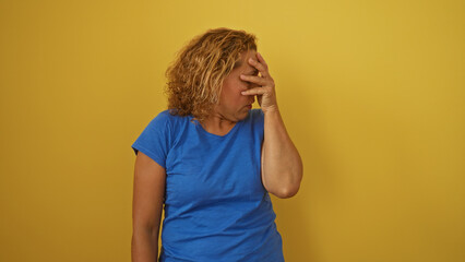 A beautiful middle-aged hispanic woman with curly hair, dressed in a blue shirt and standing against a vivid yellow wall, covers her face in an emotional and isolated pose.