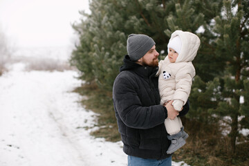 Bearded man is holding a baby dressed warmly in a white overall and hat, surrounded by snow-covered ground and evergreen trees. Joyful moment between the father and little daughter in a winter