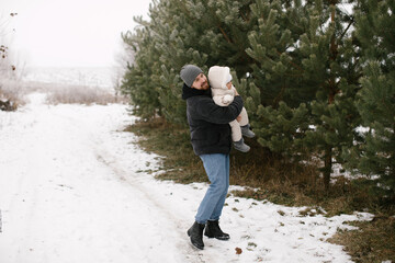 Bearded man is holding a smiling baby dressed warmly in a white overall and hat, surrounded by snow-covered ground and evergreen trees. Joyful moment between the father and little daughter in a winter