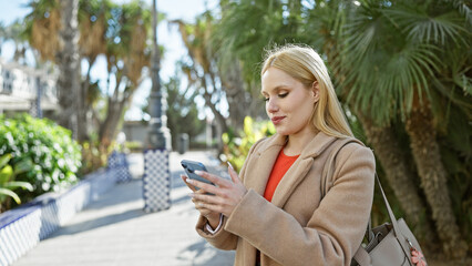 A blonde woman using smartphone outdoors in a sunny palm-lined park.