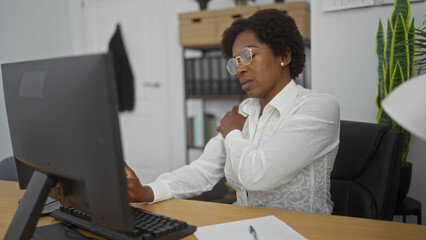 Woman working in office with computer is massaging shoulder, wearing glasses, sitting at desk, taking break, feeling discomfort, focused, workplace, adult, curly hair, indoors