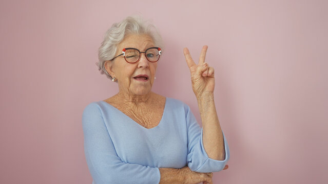 A cheerful senior woman with grey hair, wearing glasses, makes a peace sign against a pink background.